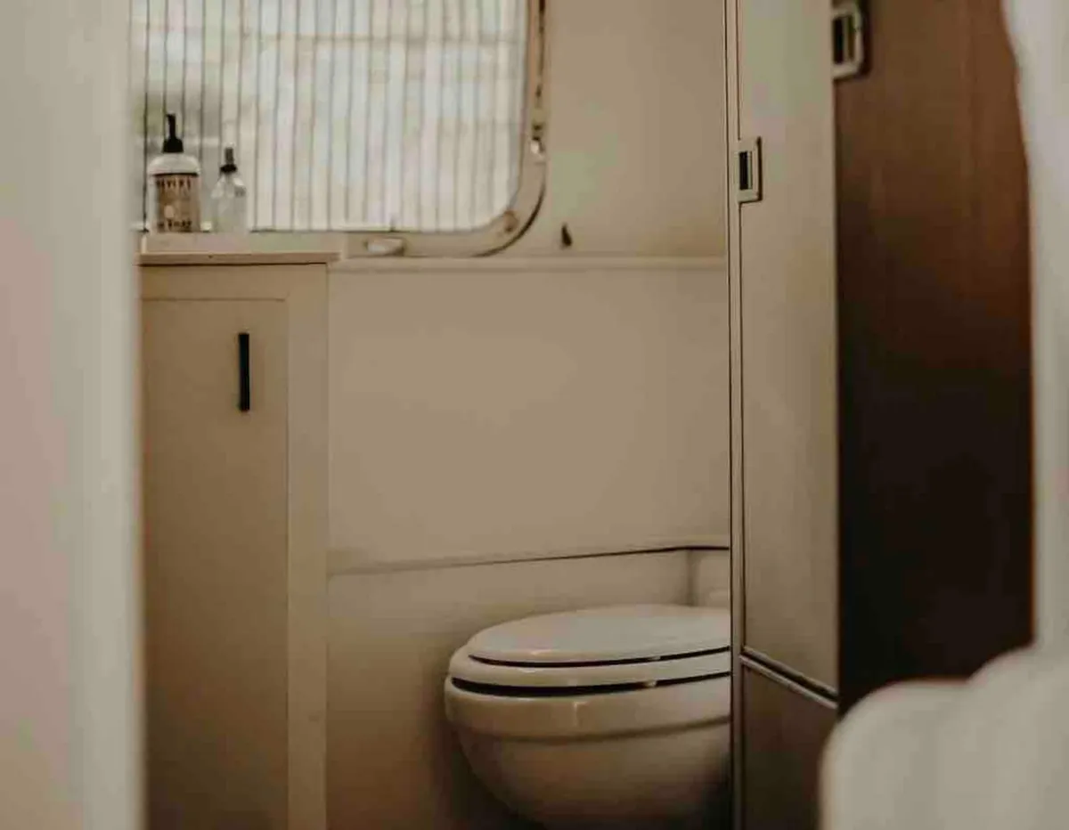 A calm, well-lit bathroom scene featuring a modern toilet, a window that brings in natural light, and a shelf with bottles that suggest thoughtful amenities. The space feels serene and clean, inviting guests to relax and refresh.