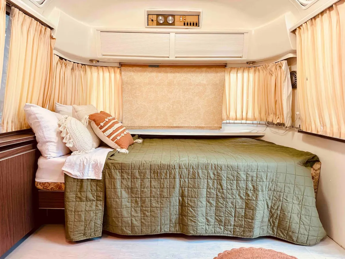 A cozy sleeping nook featuring a bed with a soft green quilt, layered decorative pillows, and curtains that frame the space. The bed is bathed in gentle light, enhancing the inviting and tranquil atmosphere, ideal for curling up with a book or relaxing after a day of adventure.
