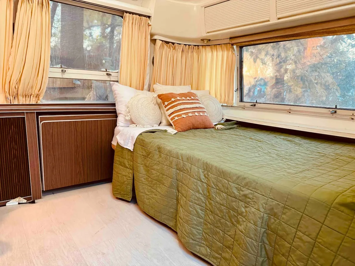 A sun-filled sleeping area with a bed dressed in a green quilt and decorative pillows, positioned near expansive windows. The scene exudes warmth and relaxation, inviting guests to wake up to natural light and enjoy peaceful mornings in a thoughtfully designed retreat.