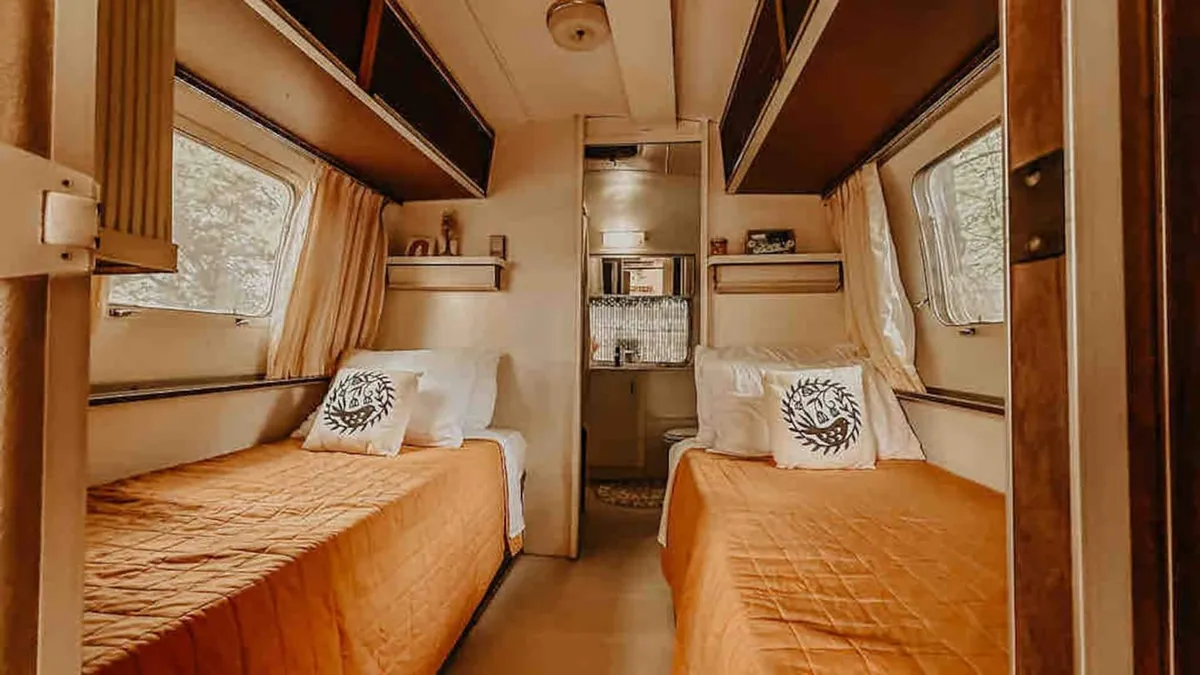Inside the Airstream, two twin beds with vibrant orange bedspreads and decorative pillows create a cozy, welcoming atmosphere. Minimal decor and wooden shelves suggest thoughtful, efficient use of space, ideal for solo travelers seeking comfort and warmth after a day of discovery.