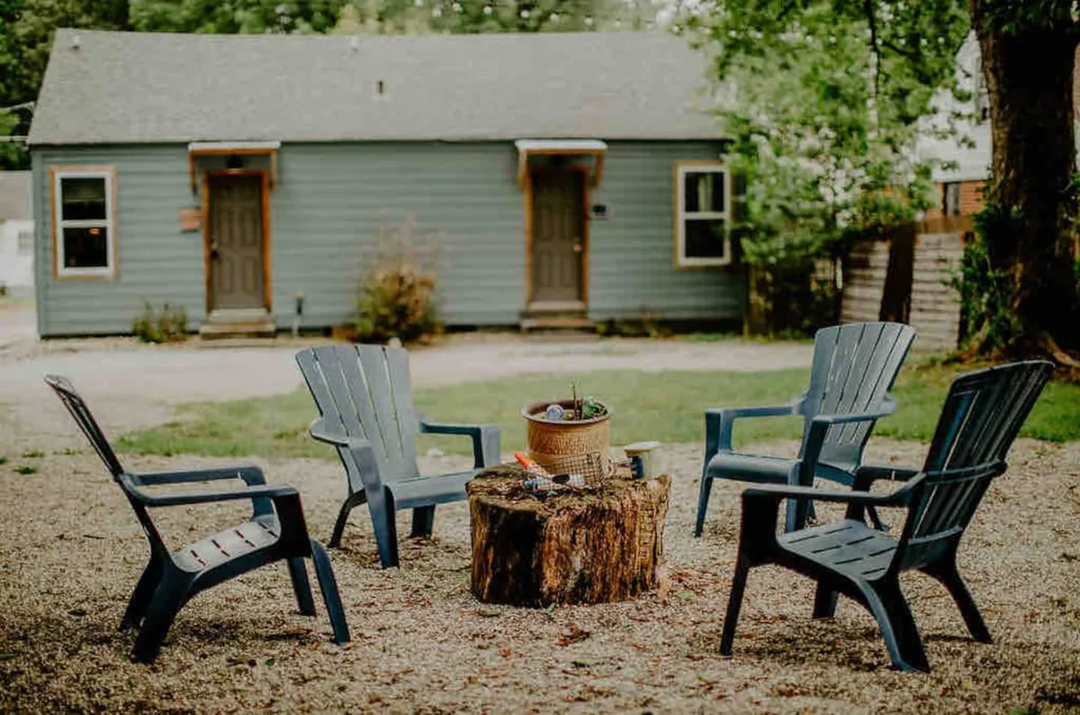 A cozy outdoor fire pit setting surrounded by Adirondack chairs and lush greenery, with a charming blue cottage in the background. The mood is inviting and nostalgic, suggesting warmth, relaxation, and shared moments under the stars.