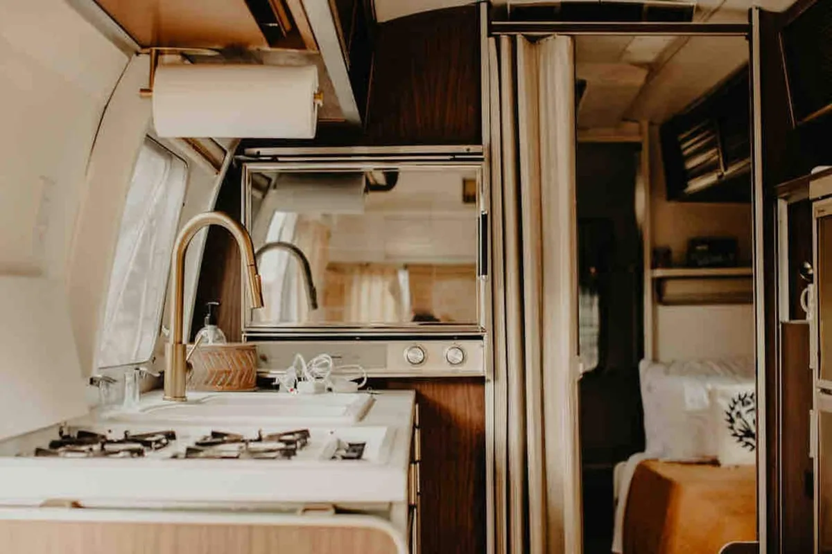 This image focuses on the kitchen sink and stove top, framed by a mirror and curtains, encapsulating a compact but functional cooking area. The retro finishes and cozy touches evoke a sense of warmth, highlighting essential kitchen amenities like the stove and sink.