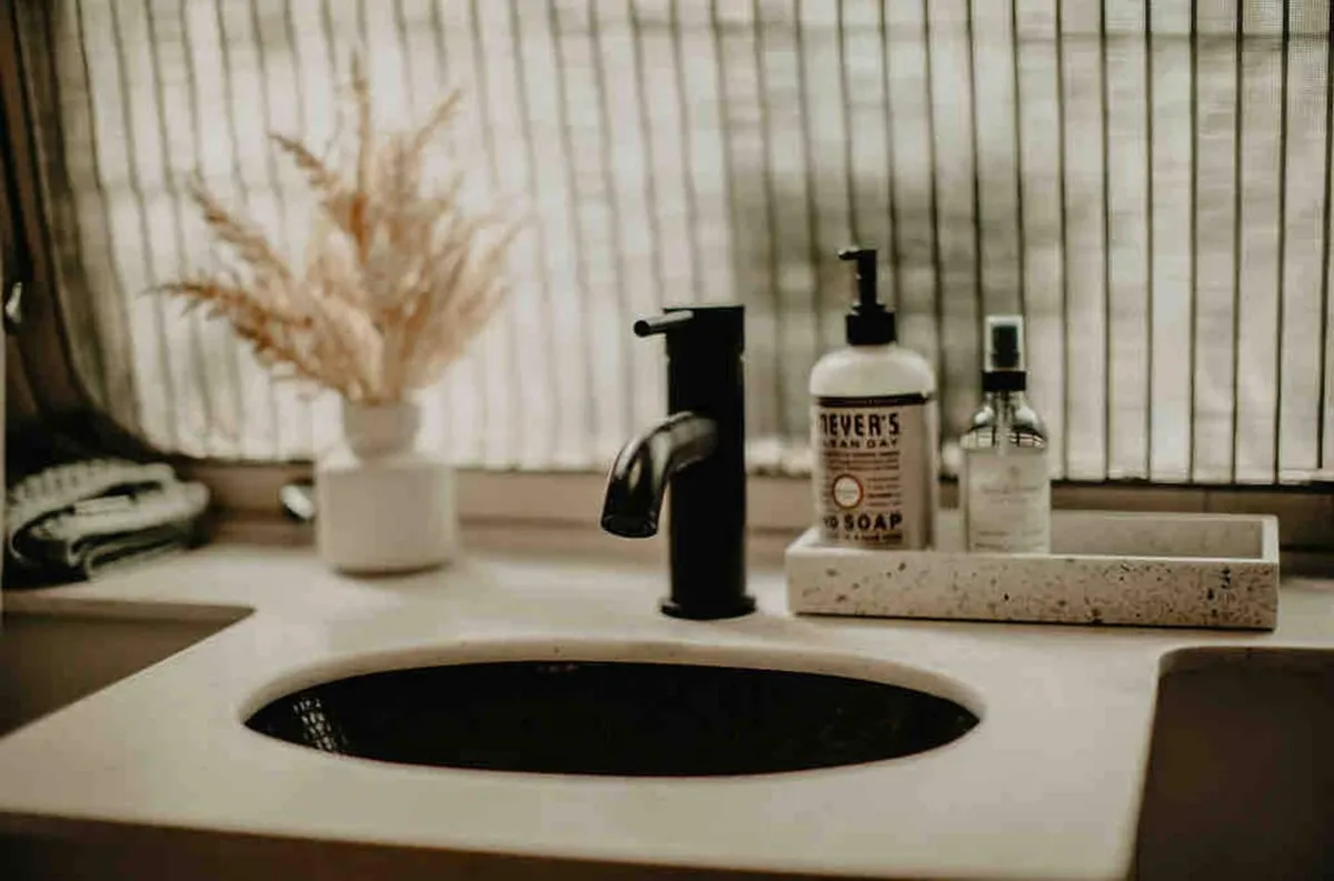 A close-up of a sleek black faucet, minimalist tray, and a small vase with dried flowers set in a calming and modern bathroom area. The scene emphasizes details that suggest both utility and design, presenting a sense of personal care and retreat.