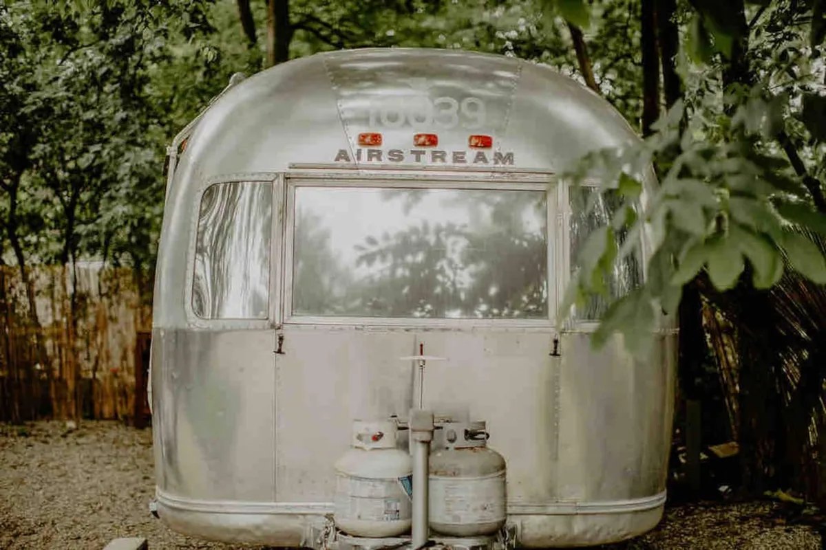 This image features an iconic Airstream trailer set amidst lush greenery. The exterior highlights the classic aluminum curves, outdoor propane tanks, and hints of a cozy porch space—suggesting opportunities for outdoor relaxation and connecting with nature.