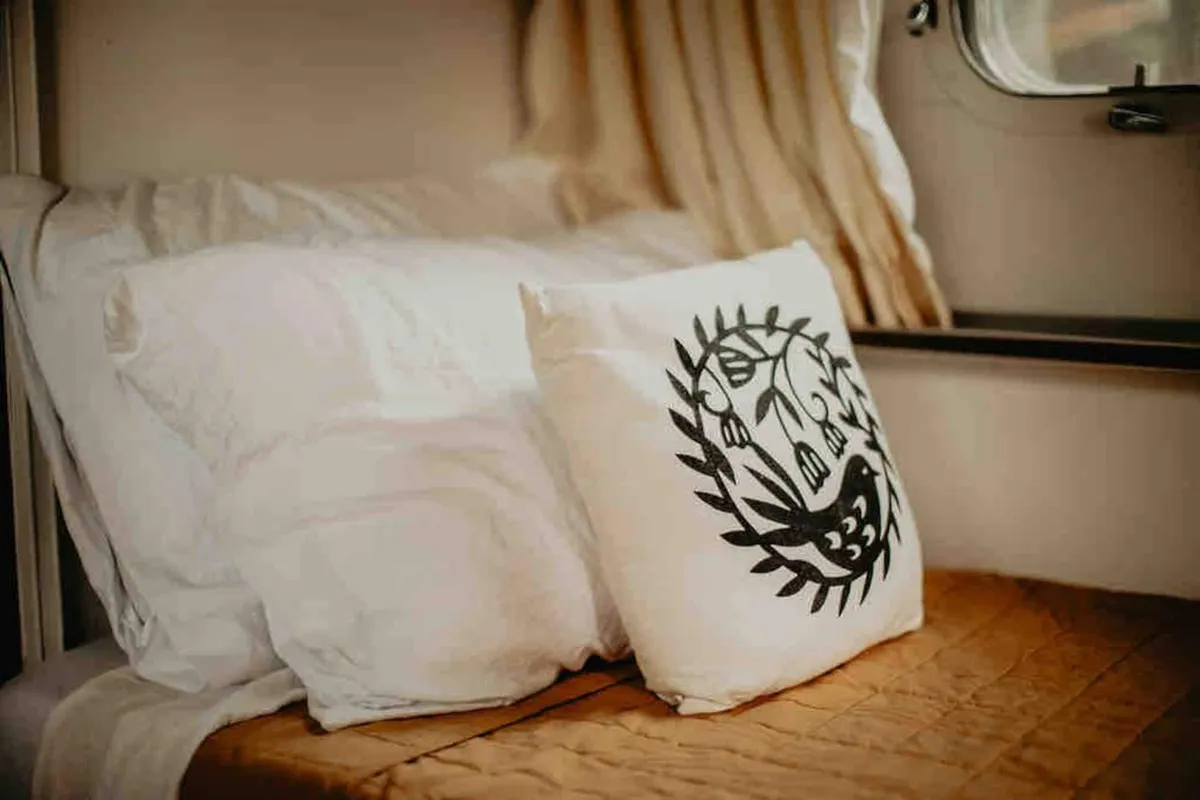 Close-up of pillows and a decorative cushion arranged on a bedspread, highlighting the plush textures and cozy layers that make the sleeping area inviting. The focus on soft details reinforces the promise of restful nights and comfortable naps.