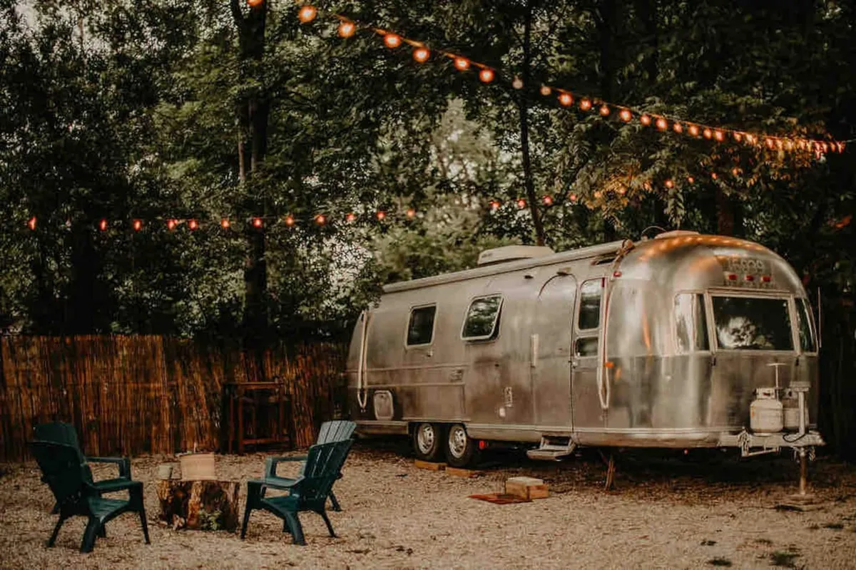 The image shows a gleaming vintage Airstream trailer parked in an urban backyard, illuminated by warm string lights. Adirondack chairs surround a glowing campfire, inviting relaxation and conversation under the stars. The scene exudes a cozy, nostalgic ambiance perfect for intimate gatherings.